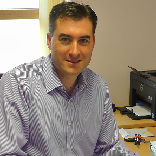 Dr Fergus Rugg-Gunn has short dark hair and is wearing a blue shirt. He is sitting at his desk and smiling at the camera.