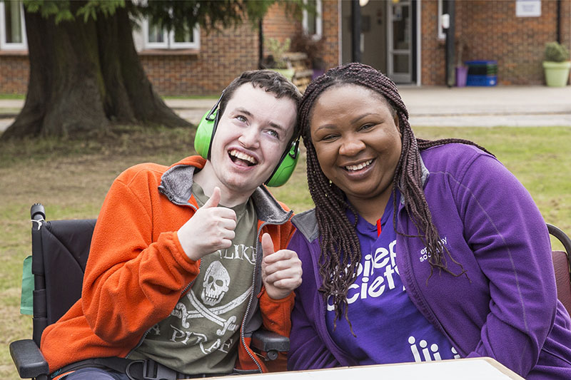 Resident at the Epilepsy Society is sitting alongside his support worker who is wearing an Epilepsy Society t-shirt. They are leaning into one another and beaming.