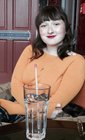 Fay has bright red lipstick and is wearing an orange jumper. She looks happy and relaxed and is enjoying a drink