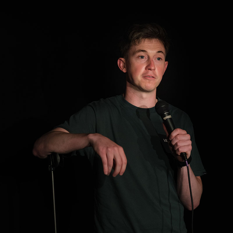 Jake is performing on stage, leaning on the mic. He looks fairly serious. The background is black