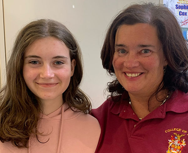 Lisa is on the right and smiling at the camera. She has a dark red polo shirt and shoulder length brown hair.  Sophia, is beside her and has long, brown hair, parted in the middle. she is smiling.