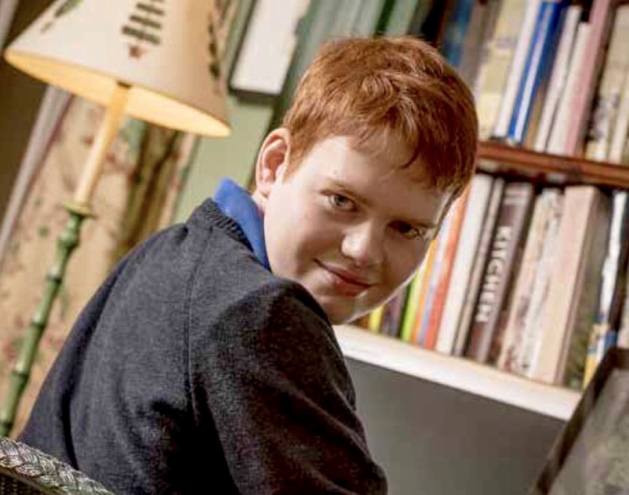 Muir is in school uniform - a grey jumper - and is grinning over his right shoulder. He is sitting at a desk, in front of a bookcase.