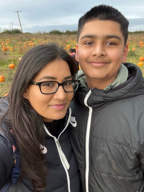 Dilan Parmar with his mum, Natasha. Dilan is wearing a dark anorak, and is smiling. Natasha has long dark hair and dark rimmed glasses. They are standing in front of a field of pumpkins