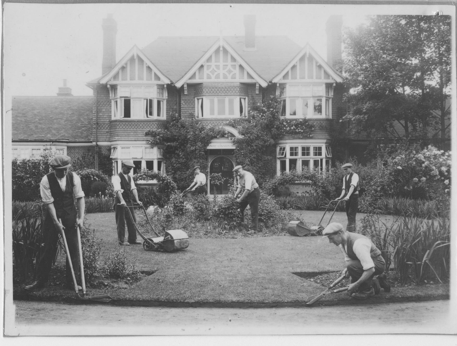 Passmore Edwards House circa 1920s with a team of gardeners mowing the grass. The Victorian-style house is covered in climbing shrubs