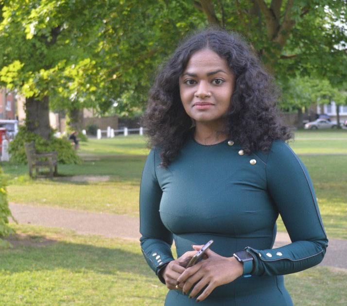 Anky has shoulder length, wavy hair. She is wearing a petrol-green dress with buttons across the shoulder and on the cuffs. she is holding her mobile.