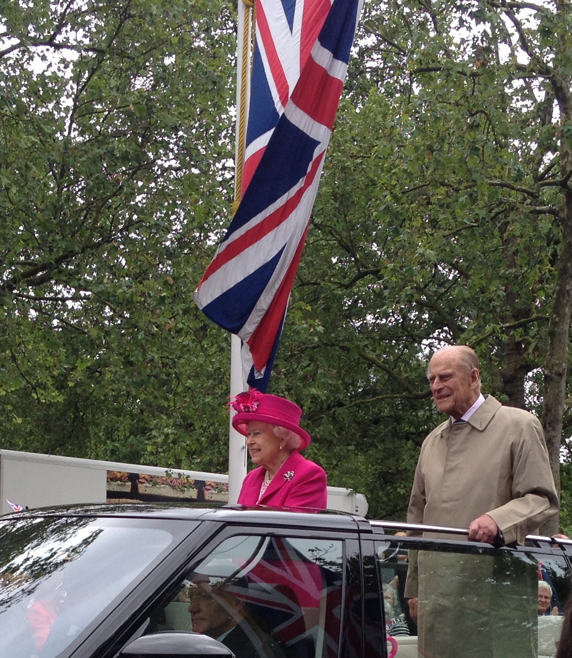 HRH Prince Philip stands alongside the Queen as they travel along the Mall in an open top car. The Queen is wearing a bright pink hat and coat. The Prince is wearing a long, beige mack.