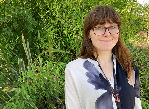 Sara Leddy is standing in front of greenery. She has long dark hair with a fringe and glasses. she is smiling