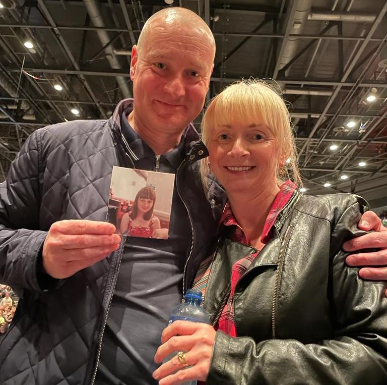 Neil and Shirley are at a concert. Neil has his arm around Shirley and he is holding a photo of Fay