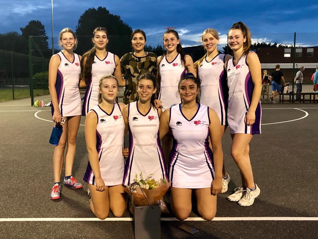 A group of girls in netball kit stand and kneel near one another outside on a netball court