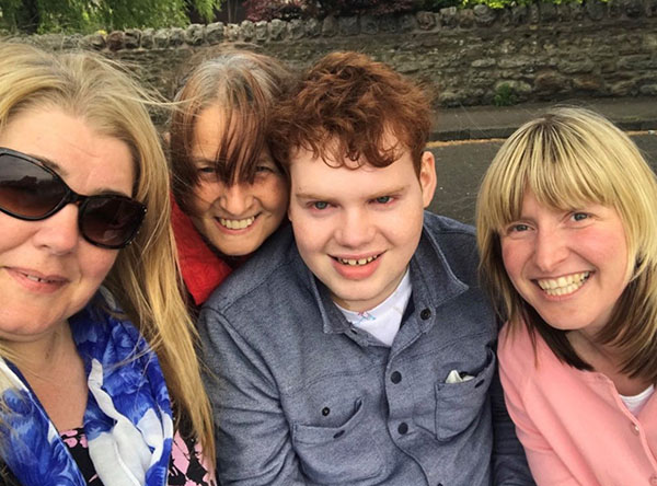 Muir is with three of his long-term carers, all are huddles round him and smiling into the camera. The sun is shining and one of the three ladies is wearing sunglasses.