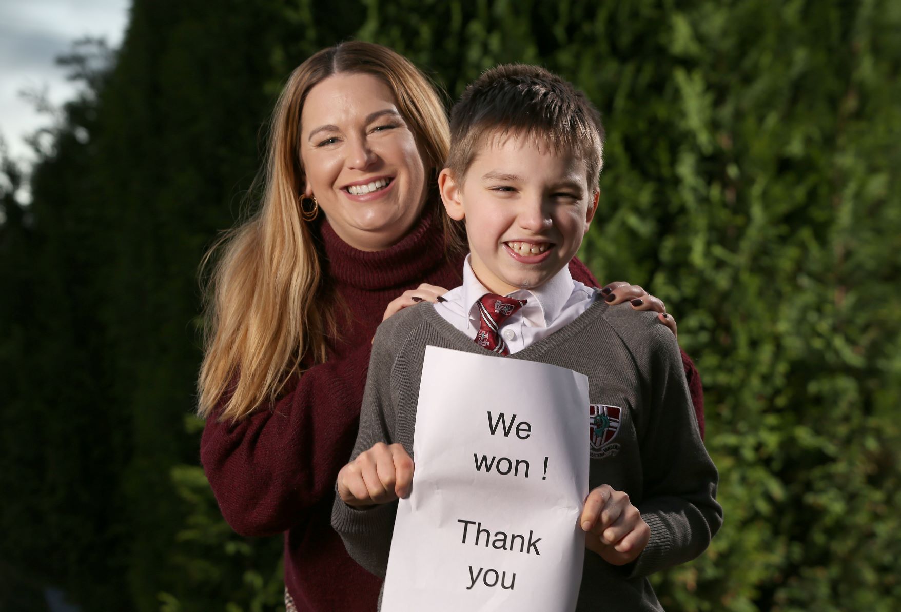 Zach, aged 11, is in grey school uniform and is holding an A4 sheet saying 'We won - thank you'. His mum, claire, with long blonde hair is standing behind him with her hands on his shoulders