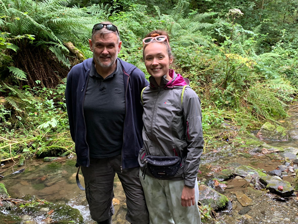 Paige Dawkins and her dad stand side by side in a stream amongst greenery smiling