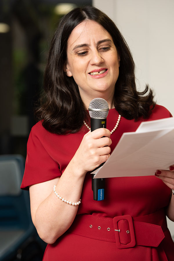 Sharon is wearing a red dress and  matching white necklace and bracelet. She has shoulder-length dark hair, parted in the middle. Sharon is talking at a mic and reading from a script.