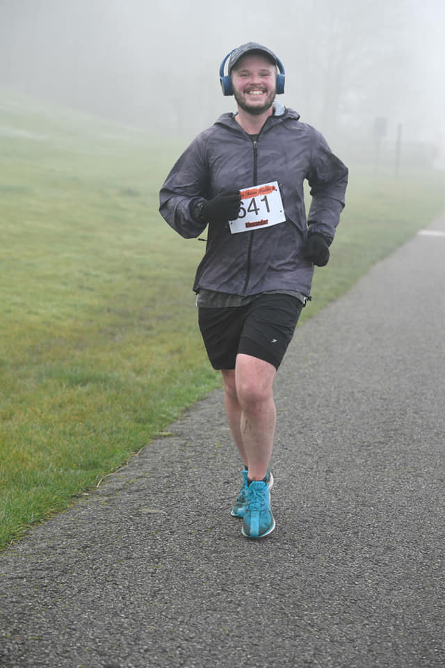Alex, AKA The Fifa Analyst, is running on pavement next to a misty field, smiling at the camera and wearing a baseball cap, headphones, a grey jacket, black shorts and blue trainers